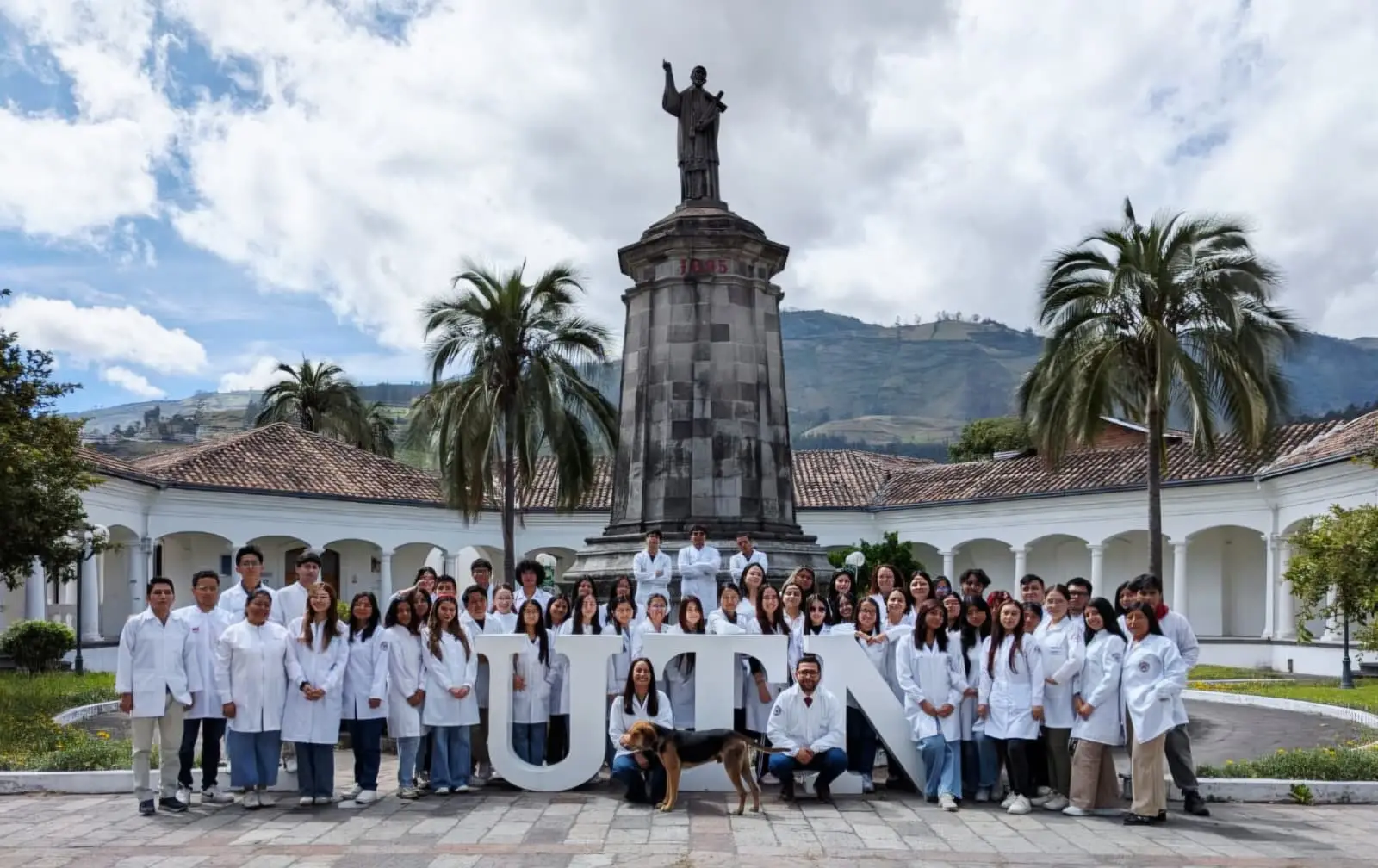 Foto di gruppo con un cane di circa 50 ricercatori dell'Università di Ibarra in Ecuador. Tutti indossano camici bianchi e sono davanti a un monumento con una statua benedicente.