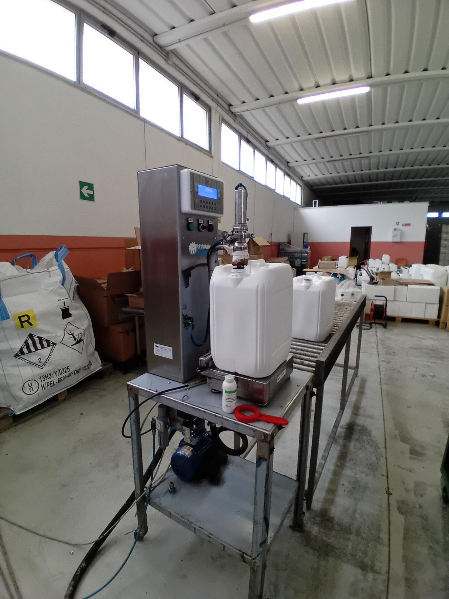 Interno di uno stabilimento produttivo di Nutrevo Q. E. In primo piano un carrello di acciaio con una pompa erogatrice mentre riempie con un liquido utilizzato in agricoltura sostenibile un fusto bianco posto su una bilancia.