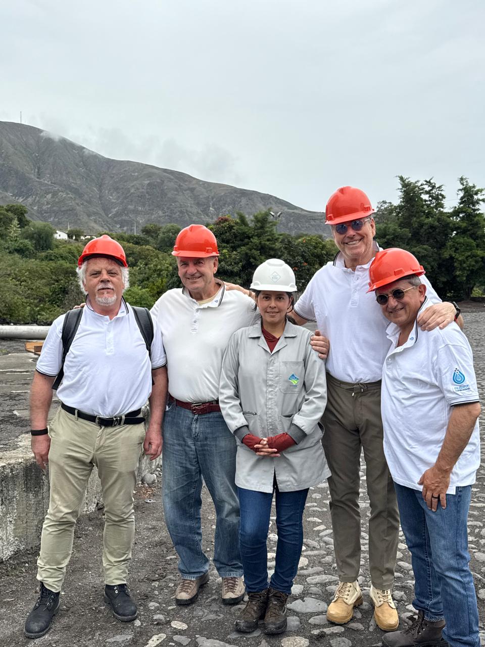 Nel corso di un progetto di produzione sostenibile in Ecuador, quattro componenti del team Nutrevo Quantic Evolution con caschi di sicurezza arancioni e una donna con casco bianco sono affiancati su uno spiazzo acciottolato. Sullo sfondo alberi, case e una montagna brulla.
